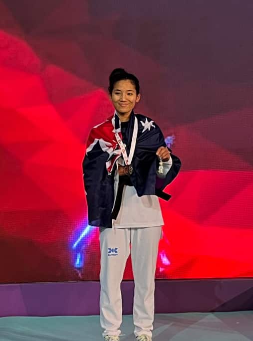 A young Nepali woman smiling on a stage. She is wearing white pants and has an Australian flag wrapped around her shoulders. 