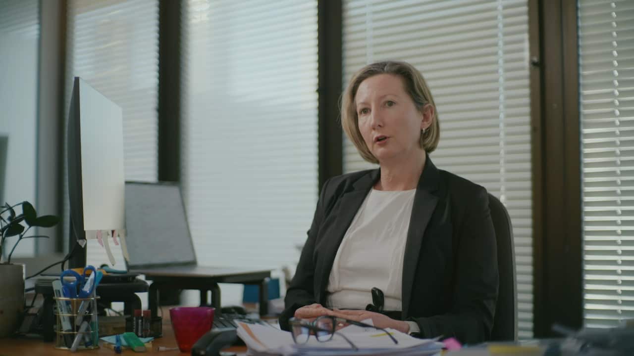 A woman with a blonde bob in a black blazer sits at a cluttered office desk with a computer screen in front of her.