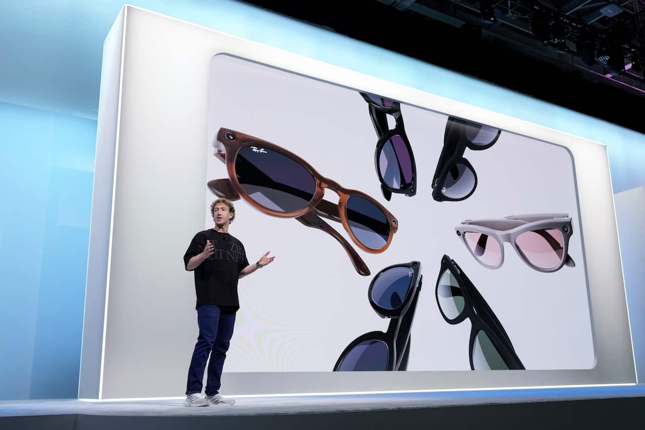 A man wearing a black T-shirt and pants stands on a stage with his arms open mid-conversation. In the background, a screen projects images of smart glasses.