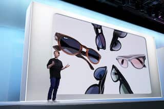 A man wearing a black T-shirt and pants stands on a stage with his arms open mid-conversation. In the background, a screen projects images of smart glasses.