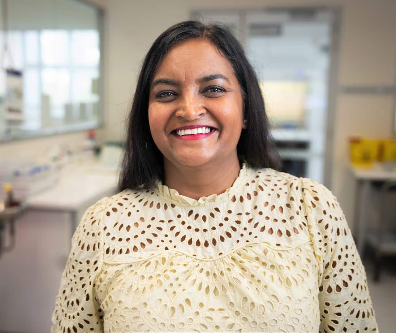 A woman with long brown hair and wearing a cream lace top stands in a science lab.