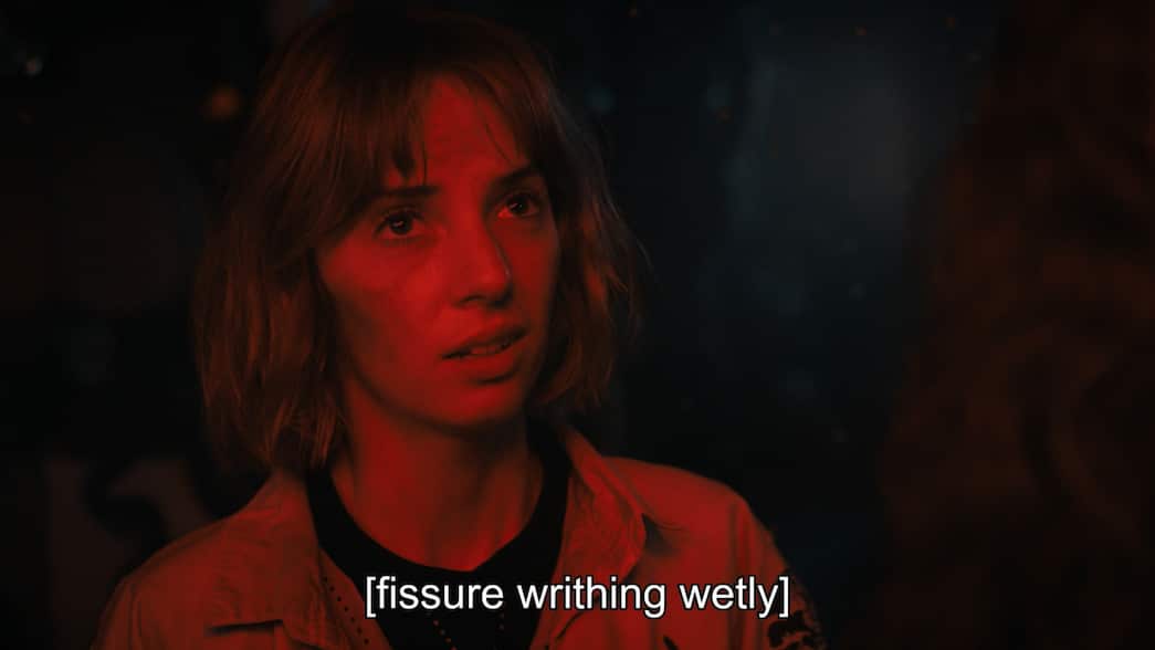 A woman with short hair is bathed in red light with closed captions reading "fisure writhing wetly"