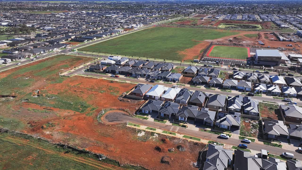 An aerial view of a suburban development shows rows of completed houses with dark roofs. In the foreground and background, large plots of undeveloped, reddish-brown land and grassy fields are visible, indicating ongoing growth.