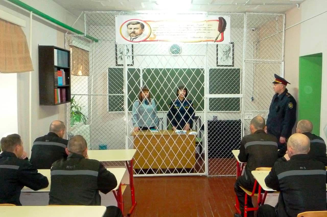 A group of prisoners sit during classes inside a prison colony. The teacher is separated from them by a metal cage.