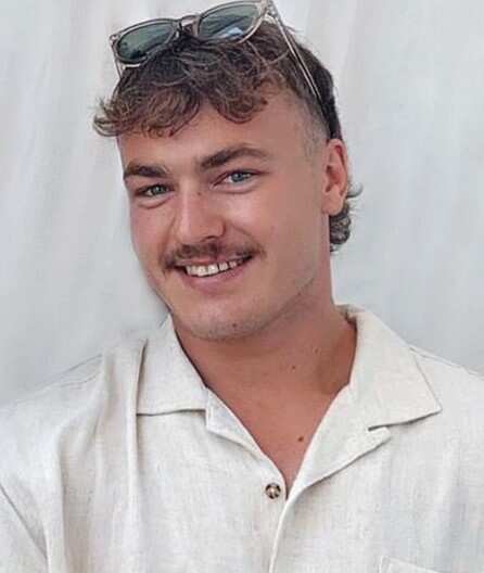 A young man with sunglasses on his head and smiling, wearing a white, collared, short sleeved shirt with tattoos visible on one arm.