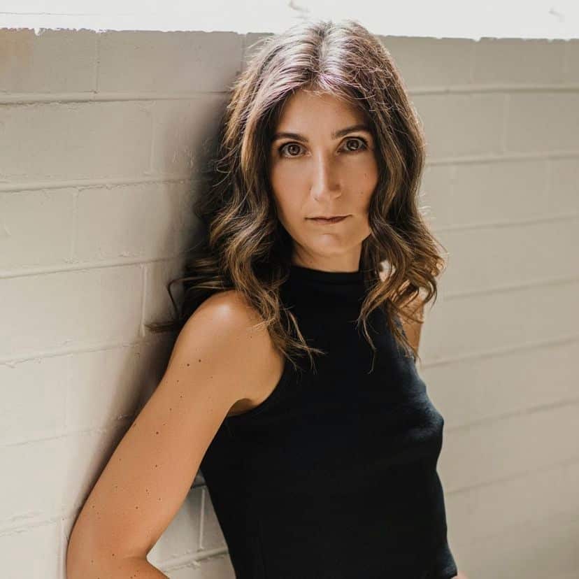 A woman in a black top leans back against the wall with a serious expression on her face.