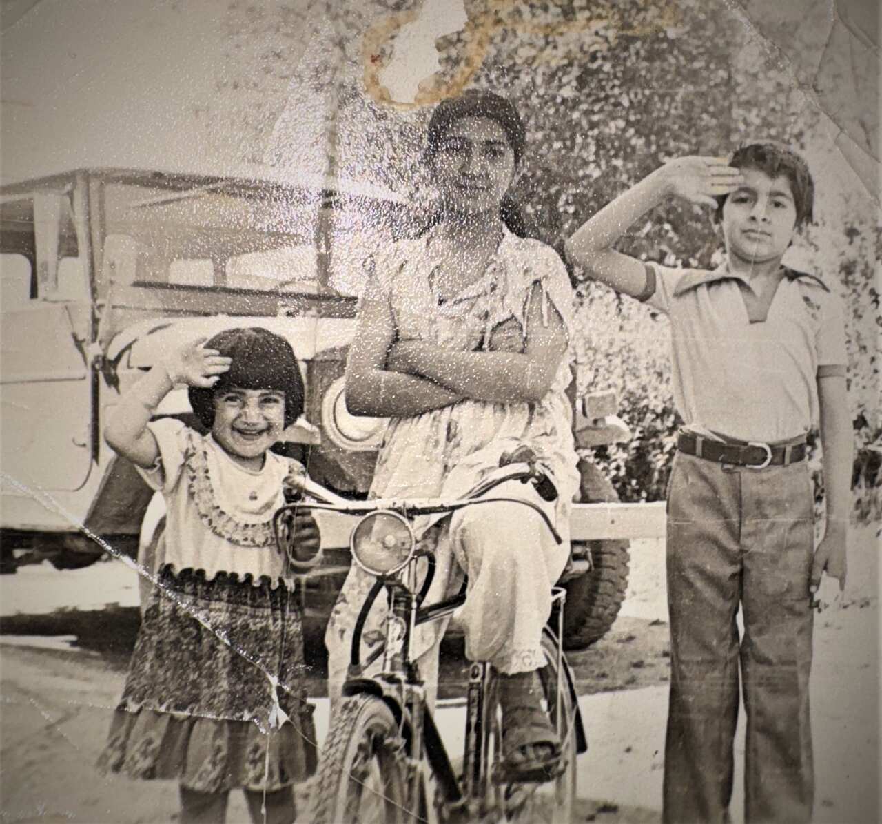 An old photo shows three young children — a girl sitting on a bicycle with her arms crossed in the middle, flanked by a younger girl and a boy standing on either side, saluting on a road next to a vehicle.