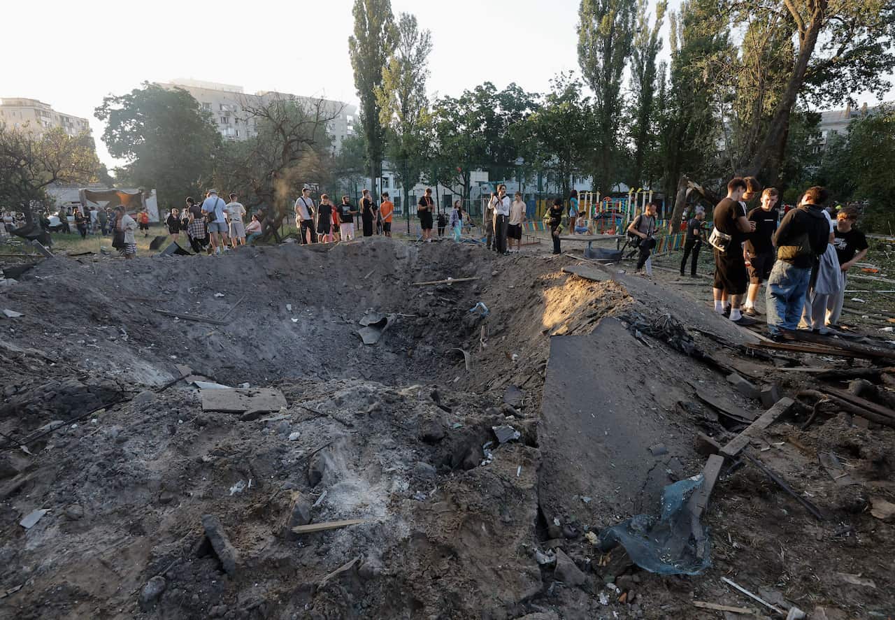 A large, deep bomb crater filled with dirt and debris sits in the foreground, surrounded by a crowd of people inspecting the aftermath near a park and residential buildings.