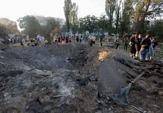 A large, deep bomb crater filled with dirt and debris sits in the foreground, surrounded by a crowd of people inspecting the aftermath near a park and residential buildings.