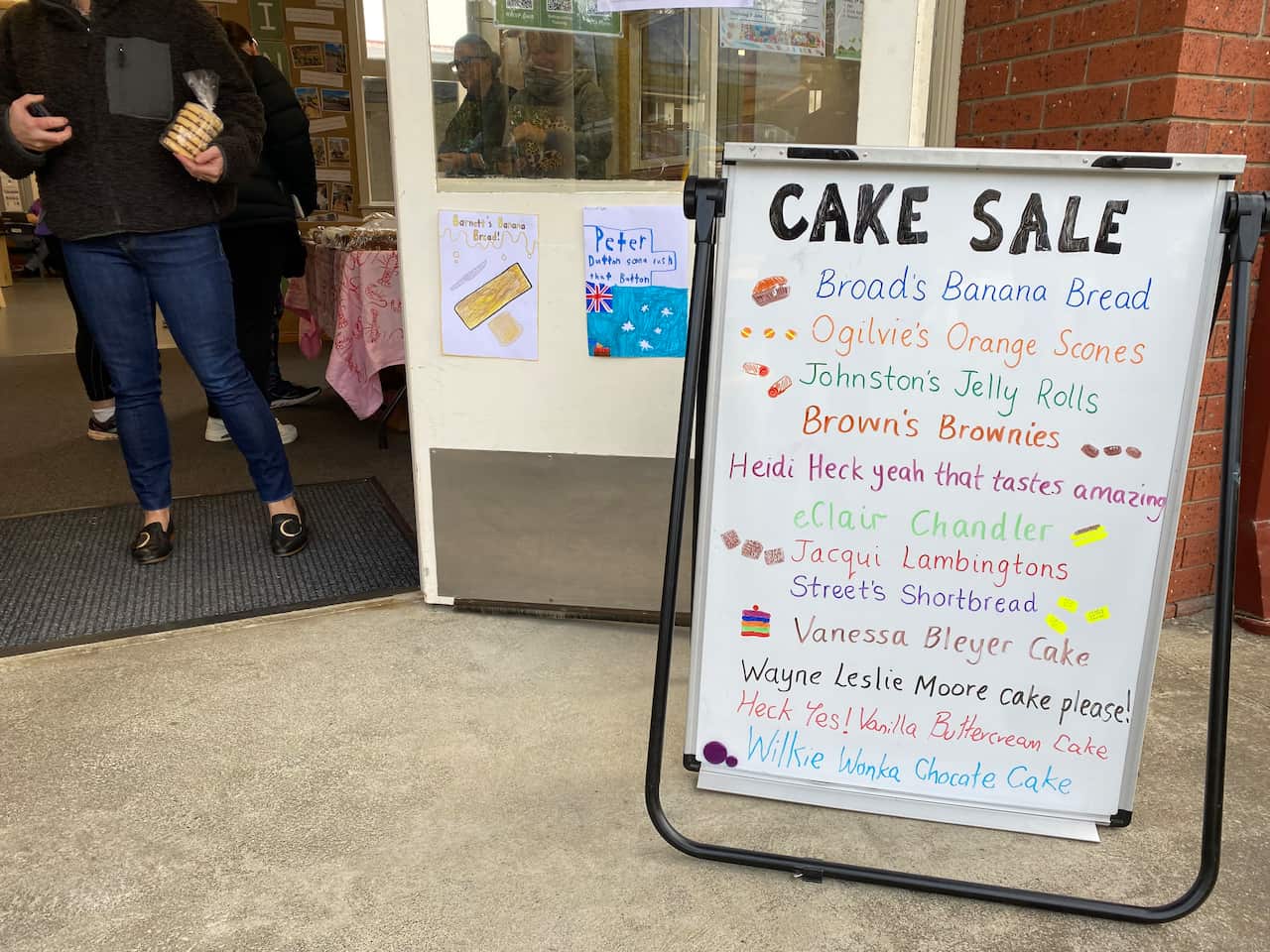 A whiteboard with novelty names of different cakes available at a cake stall set up at a polling place.