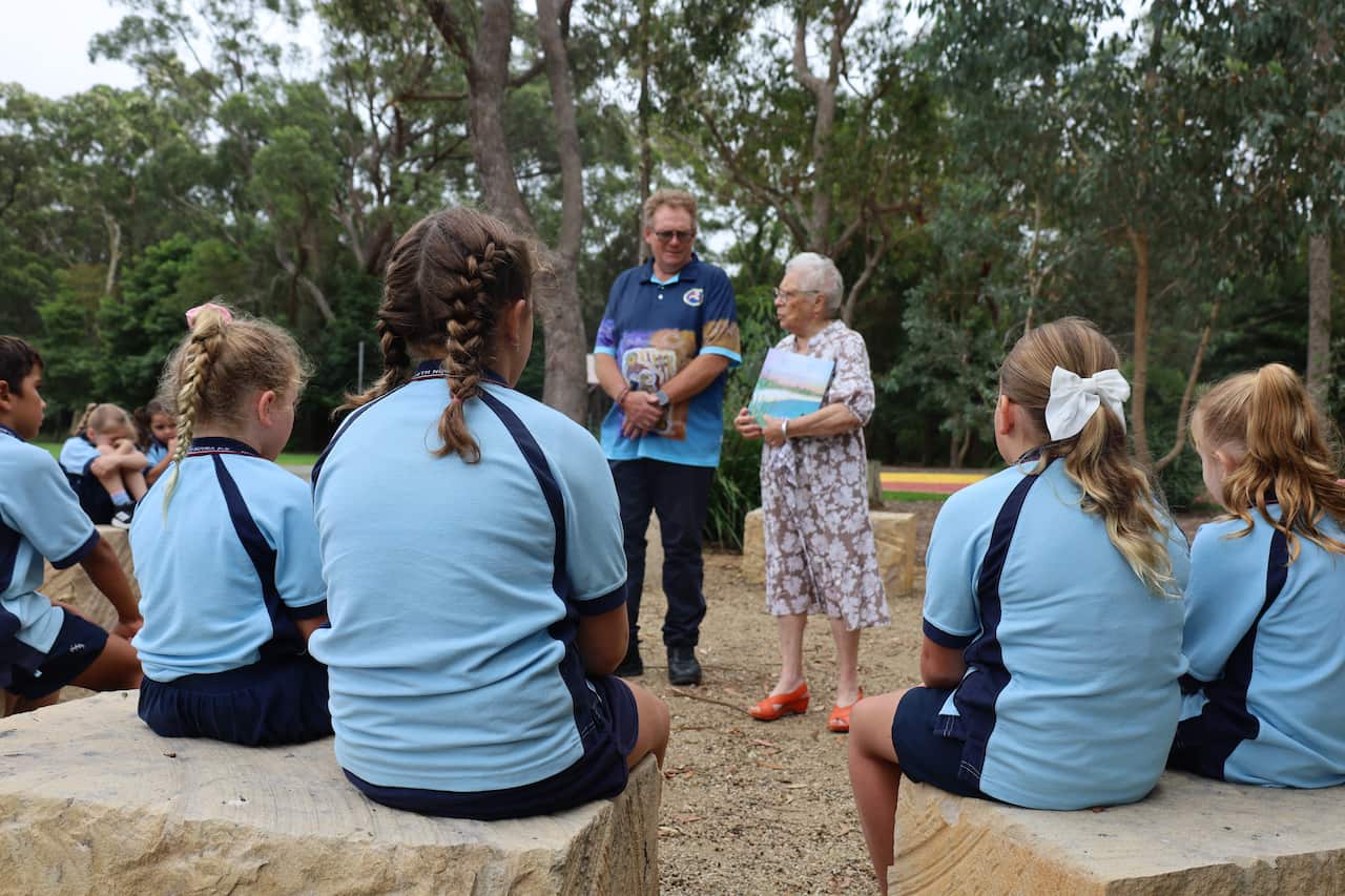 An elderly woman stands outside with a man in front of a group of young students, who sit on sandstone blocks and wear light blue polos.