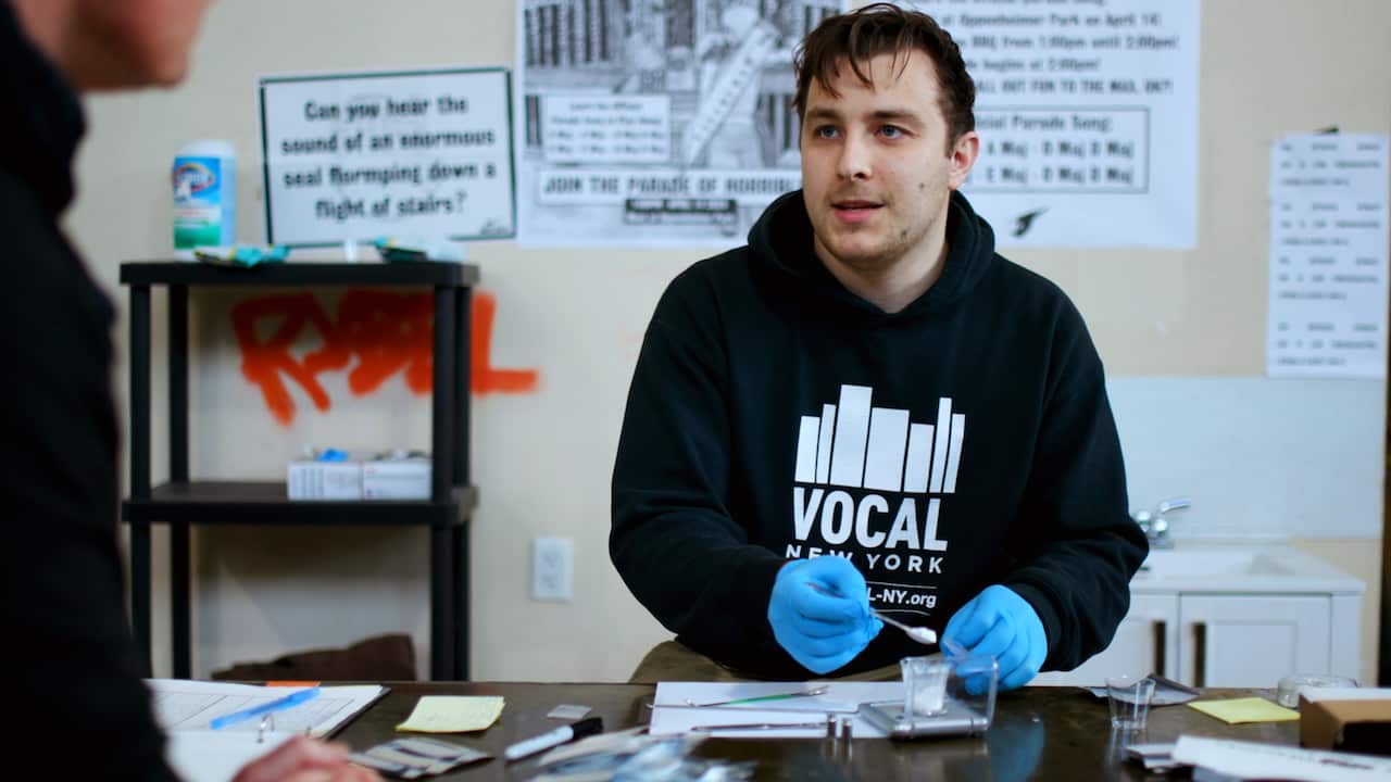 A 20-something man wearing blue medical gloves holds a spoon as he fills a plastic bag with a small dose of a powdered drug