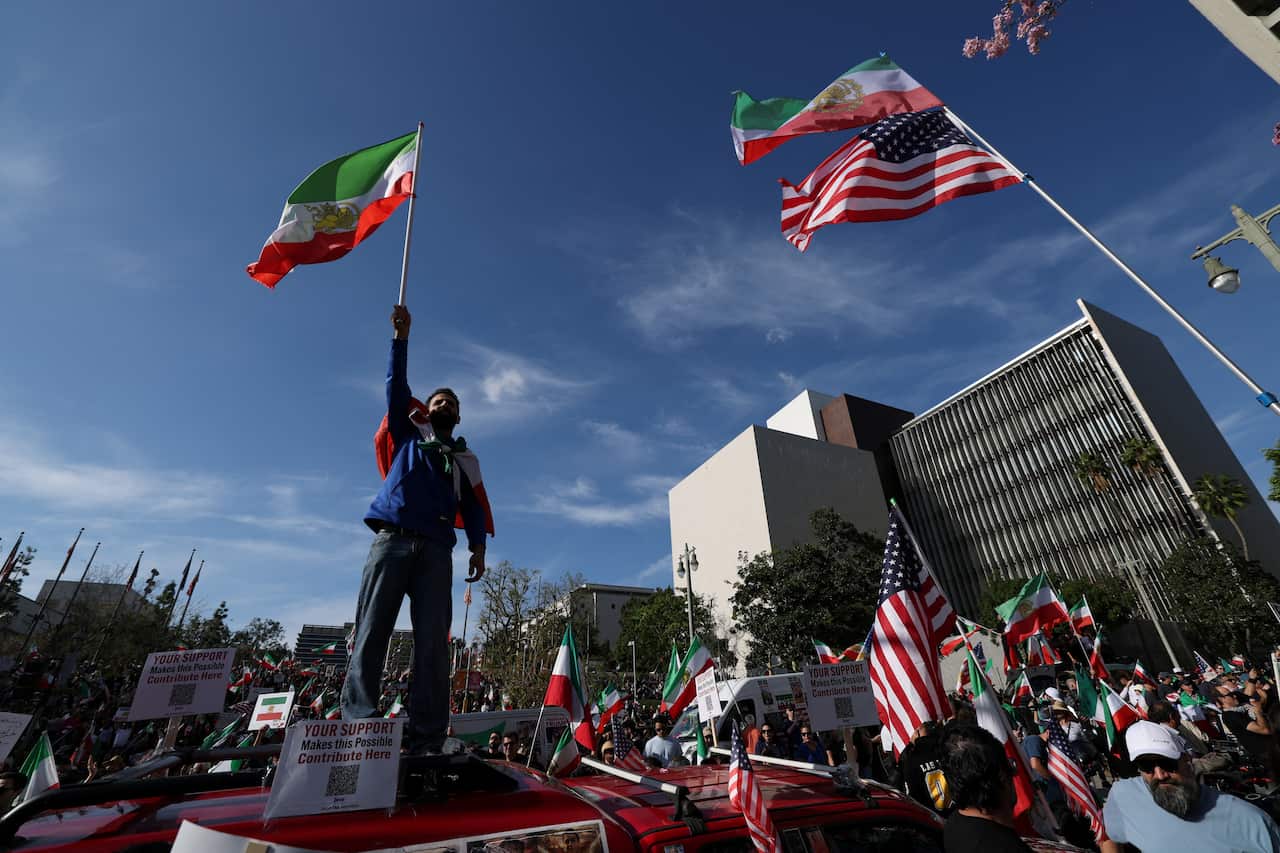 A man holding an Iranian flag high above his head stands above a crowd holding a mixture of Iranian and American flags