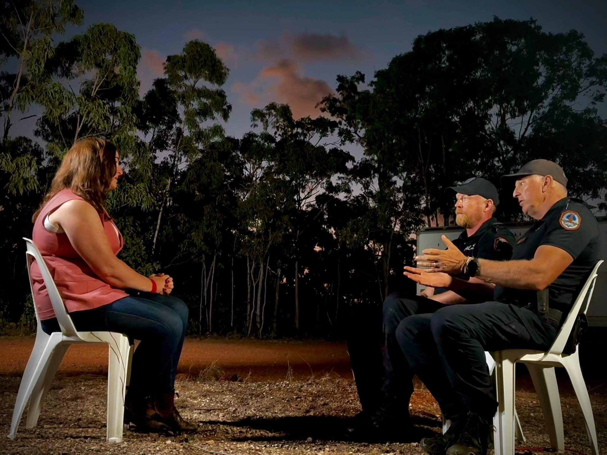 A woman in a pink shirt sitting in a plastic chair. She is speaking to two male police officer in navy uniform opposite her. They are outside and there are trees in the background. 