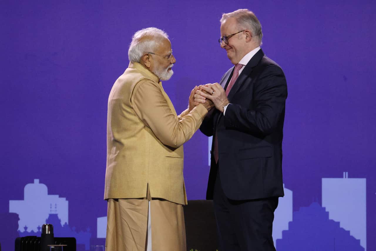 Narendra Modi,Anthony Albanese on stage holding hands in front of a purple background in the stadium.