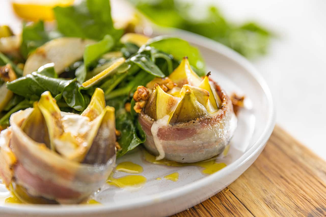 Two figs, cut open and wrapped in a band of pancetta, sit on a white plate, beside a pile of salad leaves. 