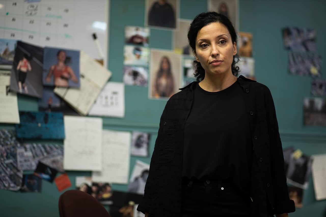 A woman stands in front of a wall mostly covered in images, and notes.