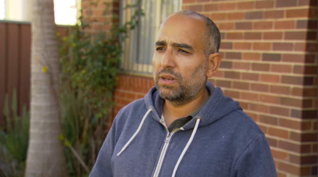A man in a blue zipper is speaking as he stands in a garden.