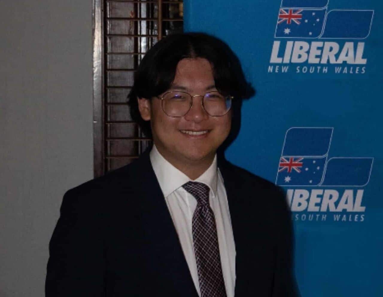 Young man in a suit standing in front of a Liberal Party NSW background.