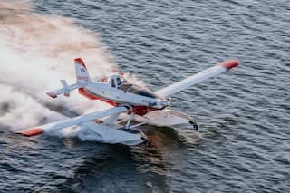 A Fire Boss plane lands on the water with its white and red body and a jet of water behind it