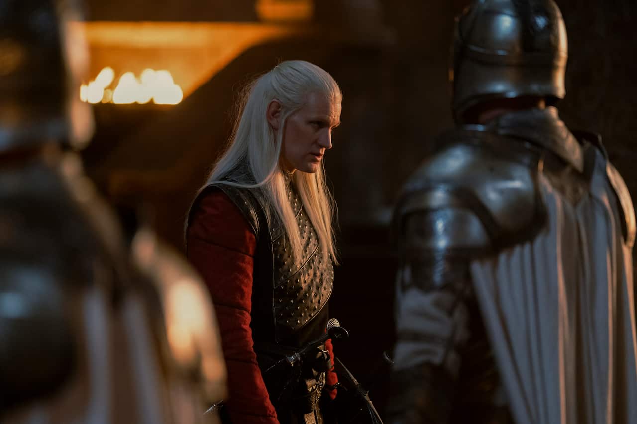 A character with long white hair looking serious in front of armoured men. 