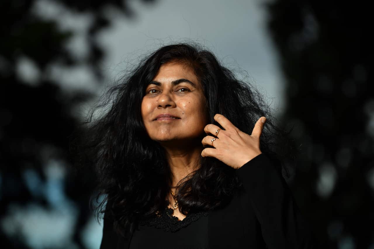 A woman wearing a black top stands with her left hand pulling back her long black hair.