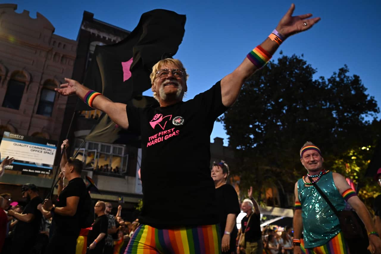 A man in a black round-neck t-shirt and rainbow-coloured shorts smiles with his arms spread wide as he marches alongside others.