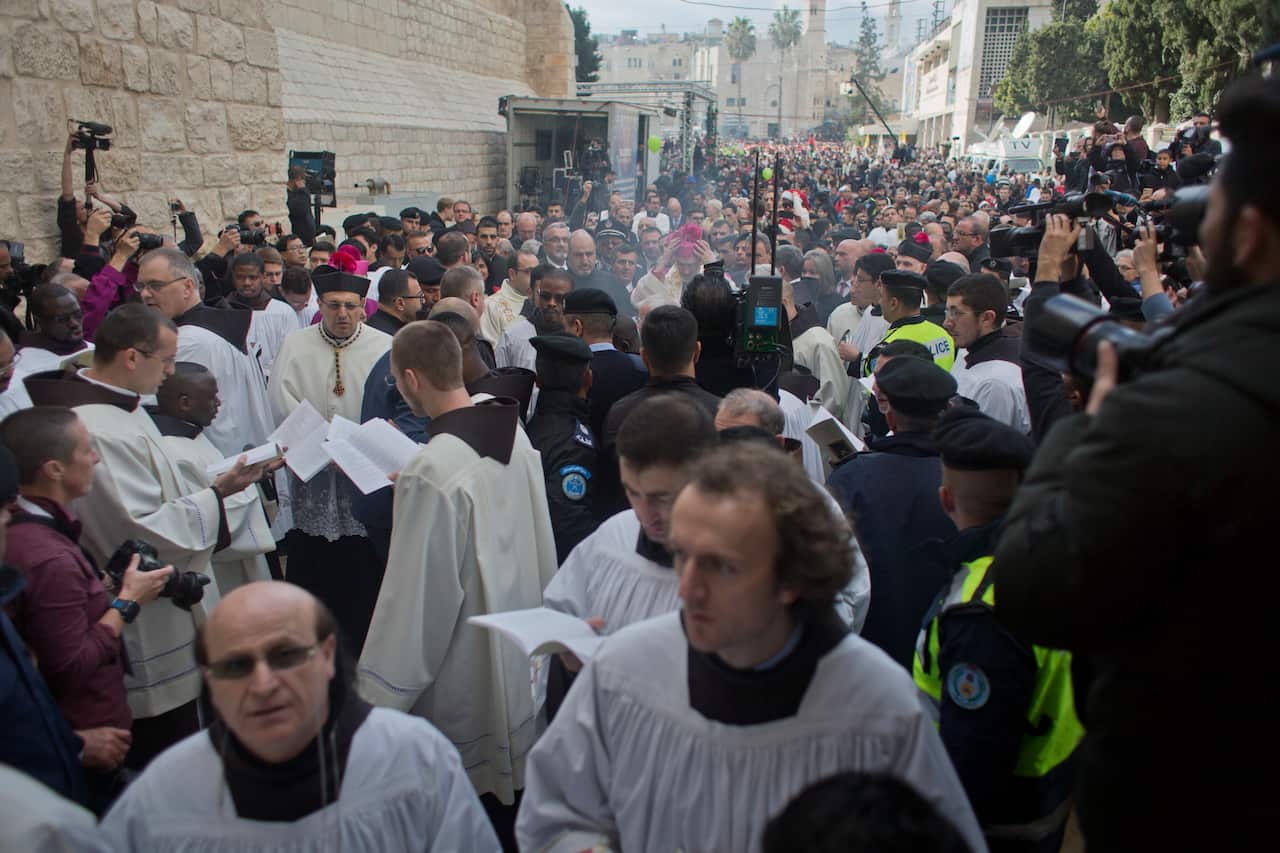 A large crowd of religious leaders, security personnel, and onlookers gather in a narrow street in Bethlehem during a Christmas procession.