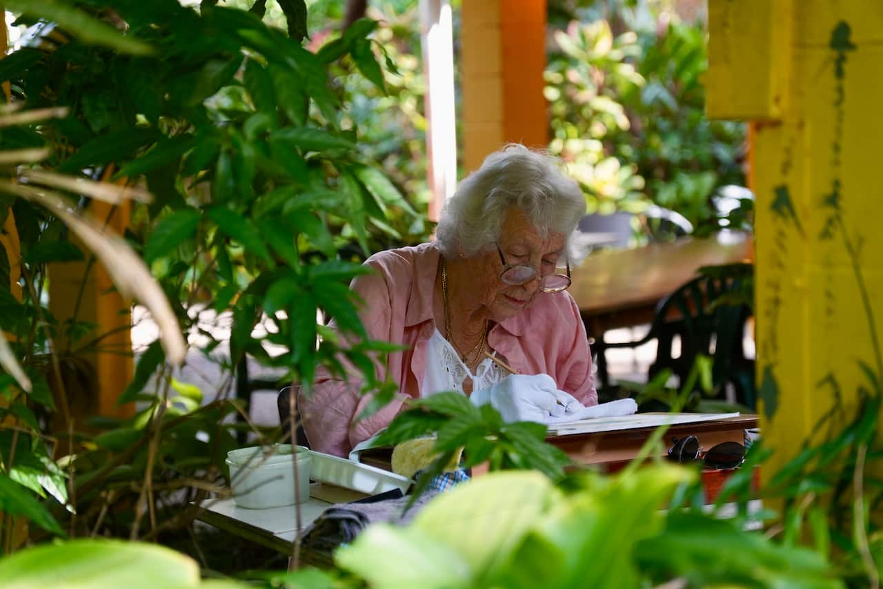 An older woman in a pink shirt worn over a white top is drawing as she sits at a table on a verandah. She is surrounded by ferns and trees.