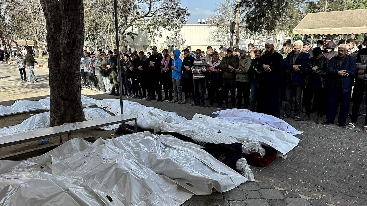 Groups of people dressed mostly in black stand in front of rows of body bags.