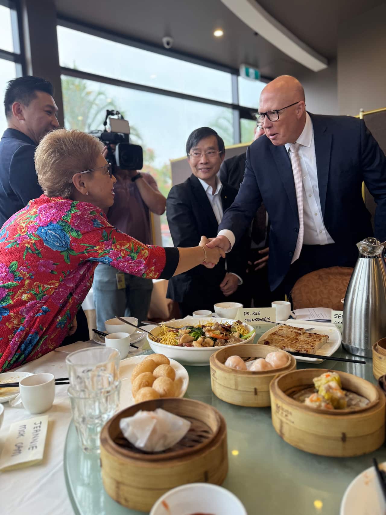 Peter Dutton shakes a woman's hand across a table full of yum cha dishes. 
