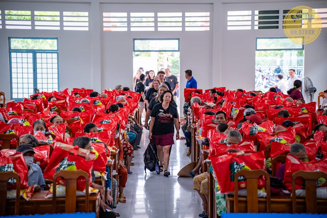A large community hall filled with Vietnamese people, each with a large red bag in front of them, with several people walking down the centre aisle.