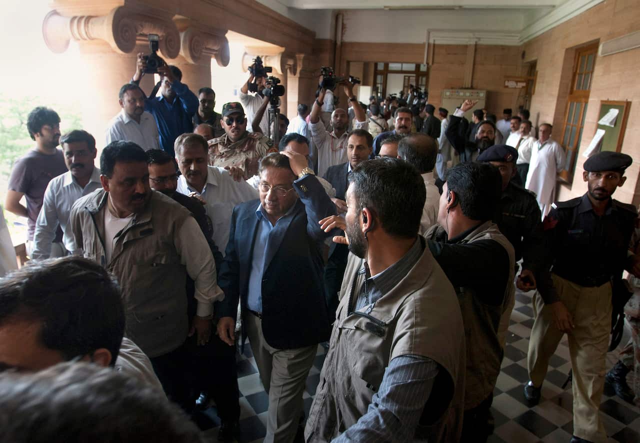 A man in a suit jacket walks with his hand on his head surrounded by mean in khaki vests. 