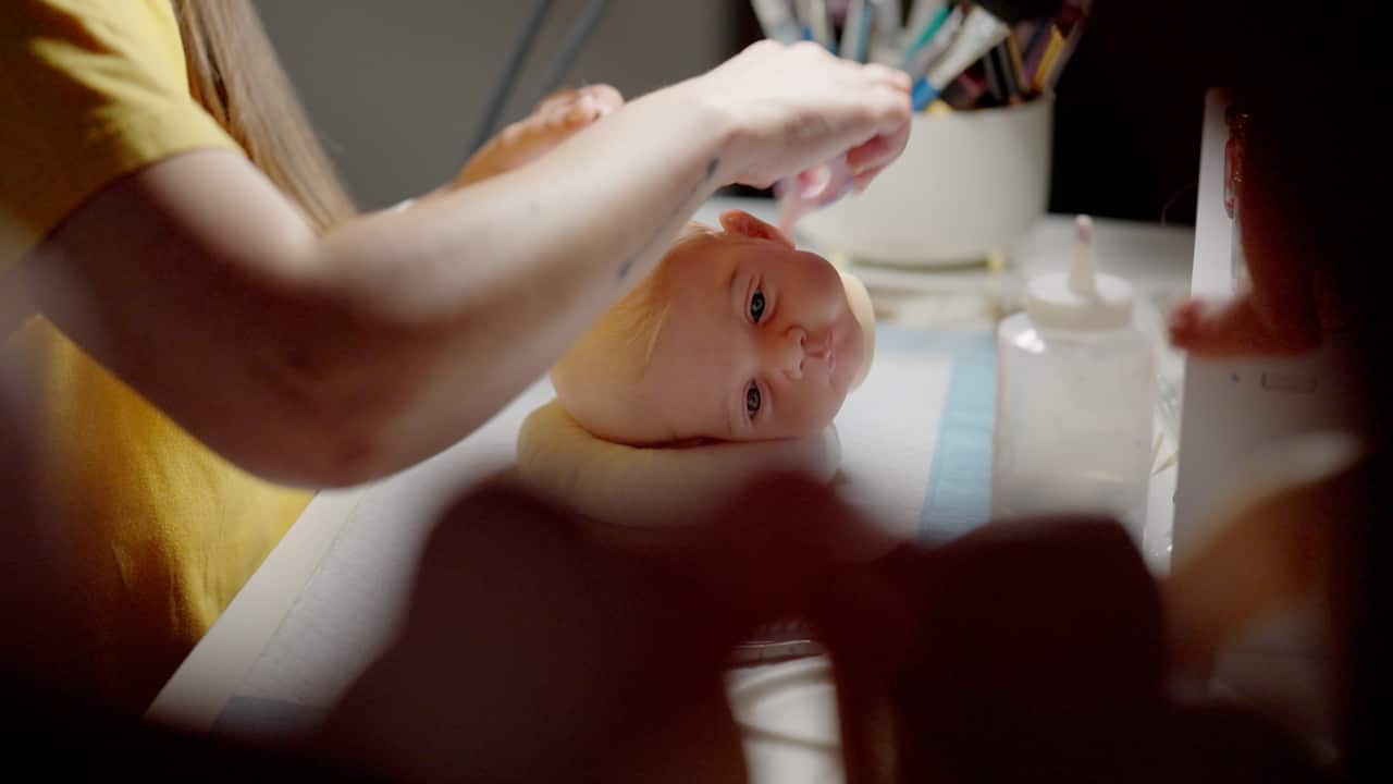 A woman works on a lifelike doll head. The head rests on a cushion, with a bottle of clear liquid and a pot full of paint tubes beside it