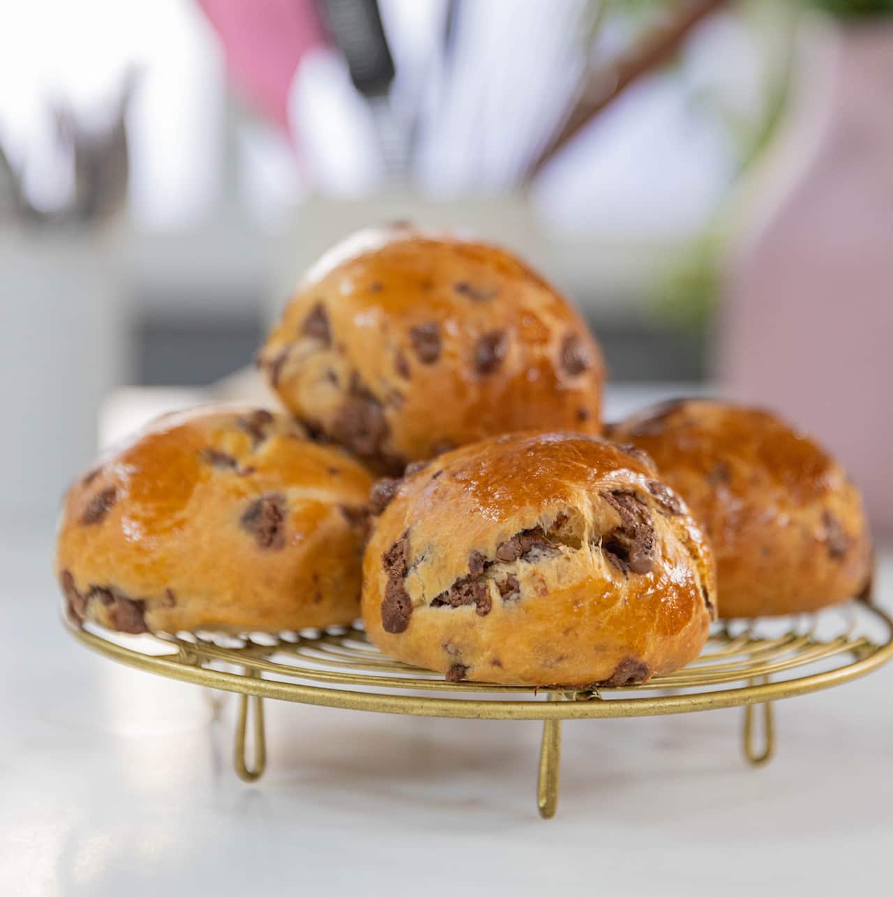 Four glossy, chocolate studded buns sit on a round, gold rack. 