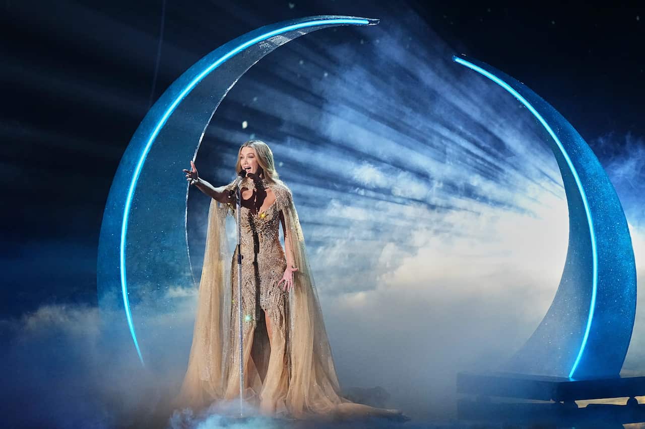 Singer Delta Goodrem, a woman with long blonde hair, in a gold dress and flowing floor-length gold scarf, performs onstage in front of a large half moon. Her arm is outstretched as she sings. The moon is backlit and the stage is filled with smoke.