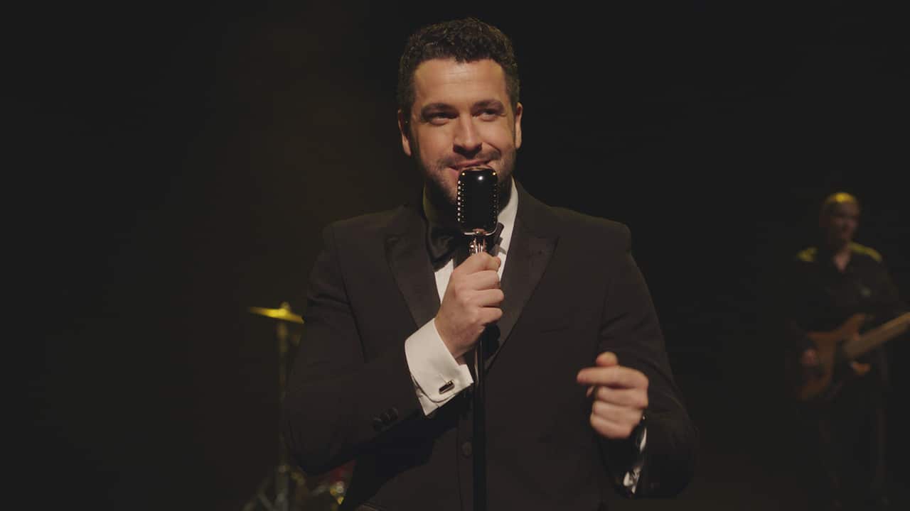 A smiling man in a suit stands holding a microphone. A musician can be seen out of focus behind him.