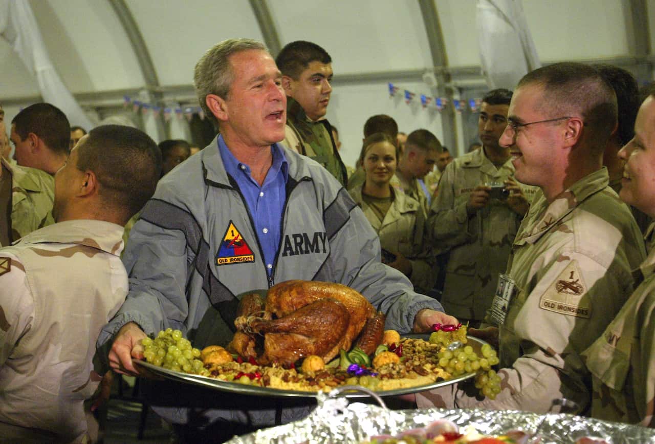 George W Bush carrying a platter with a roast turkey and fixings as soldiers watch on.