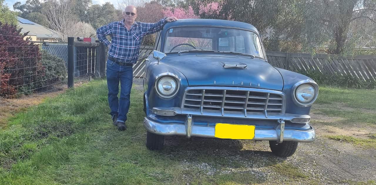 A man in a checked shirt and sunglasses stands next to his old-model blue car.