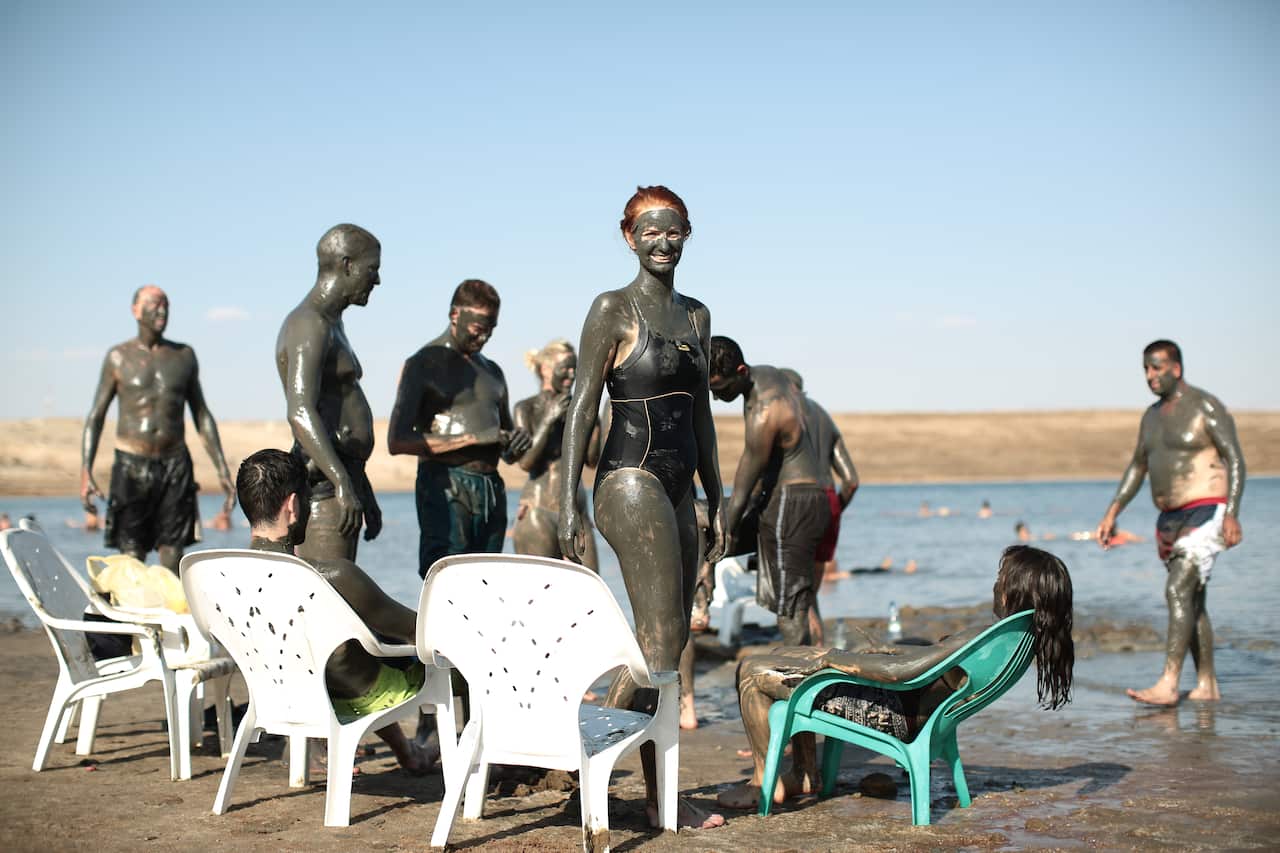 A group of bathers lathered in dark mud gathered around several plastic chairs on the edge of a body of water.
