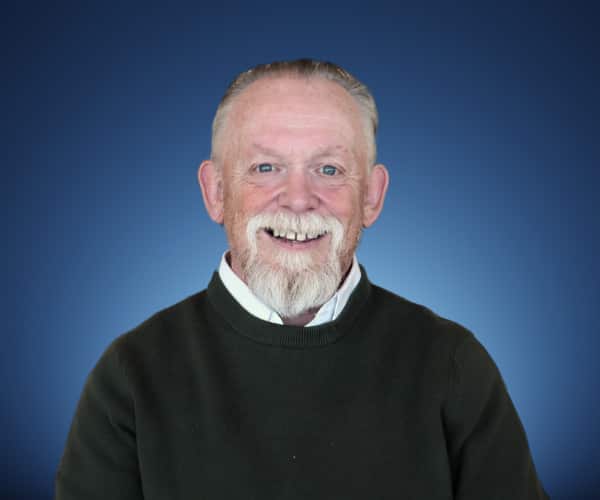 A man with a beard smiles as he sits in front of a blue background. 