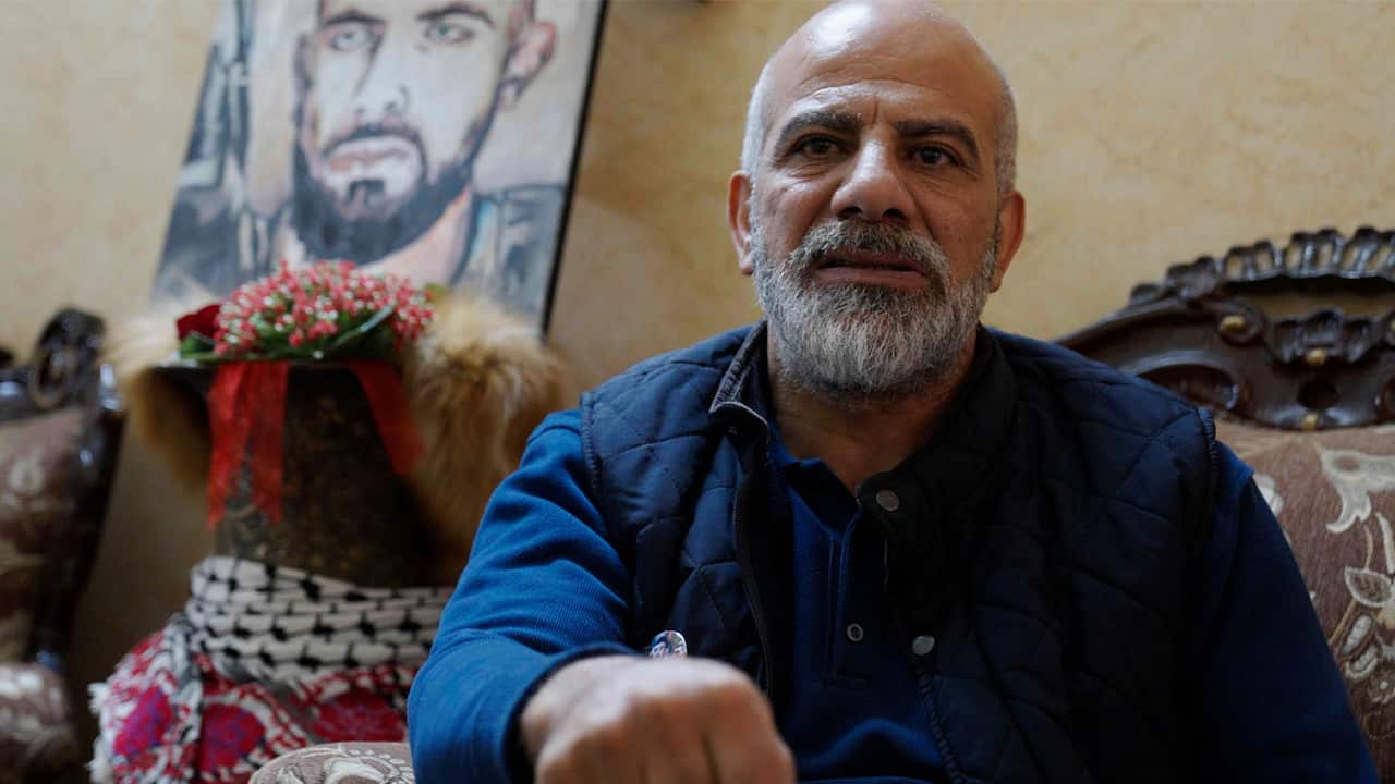 An elderly man with a grey beard and wearing a blue shirt and a vest sitting on the couch with a portrait of a young man behind 