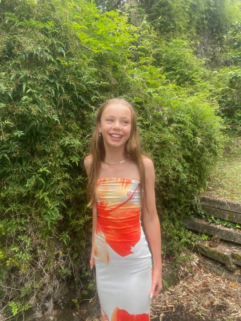 A young girl in a red and white dress is smiling with the backdrop of a bush