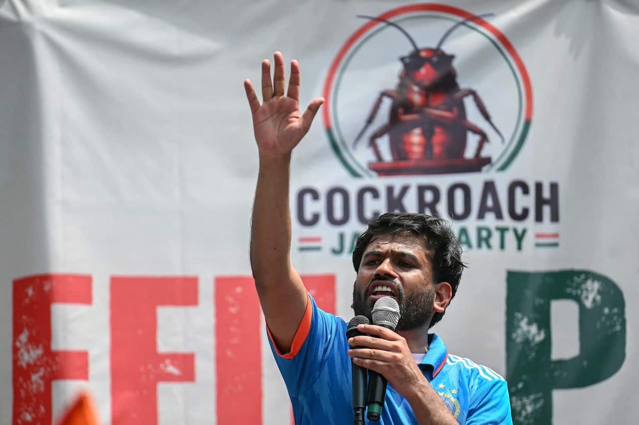 A man speaking into two microphones and raising his hand in the air. A banner with a cockroach and "Cockroach Janta Party" is behind him.