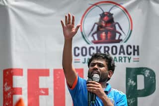 A man speaking into two microphones and raising his hand in the air. A banner with a cockroach and "Cockroach Janta Party" is behind him.