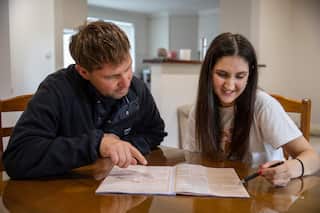 A man and woman sit at a desk looking at an orange piece of paper. The man points at a questions and the woman holds a pen.