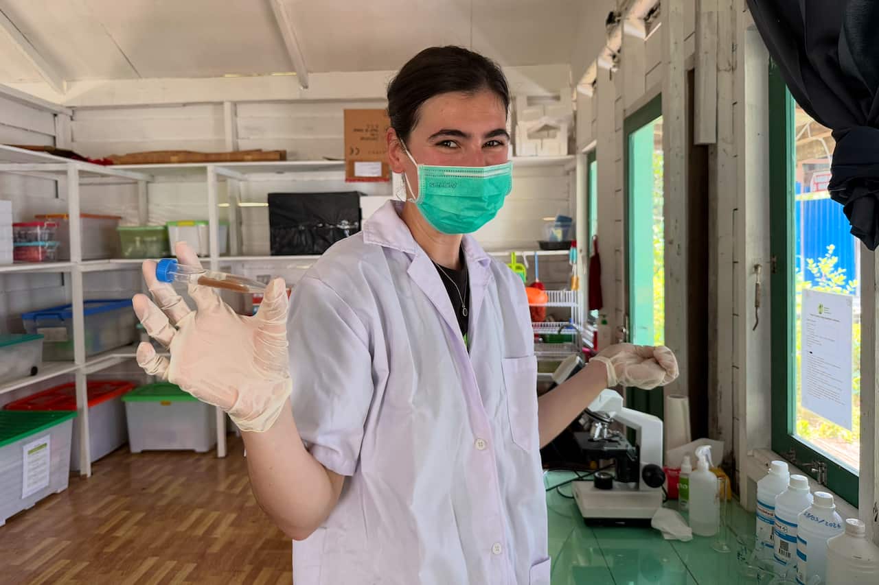 A woman in a white coat and wearing a light green face mask holds up a vial in her right hand