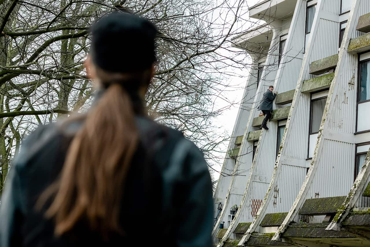 A woman in a dark cap and jacket, back to camera, stands looking at man clinging to a balcony part-way up a tall building. 