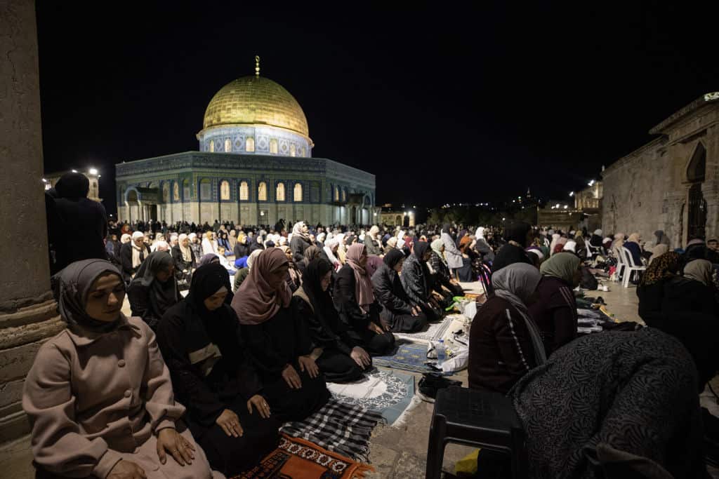 People kneeling in prayer outside at night. The Al-Aqsa Mosque is in the background.