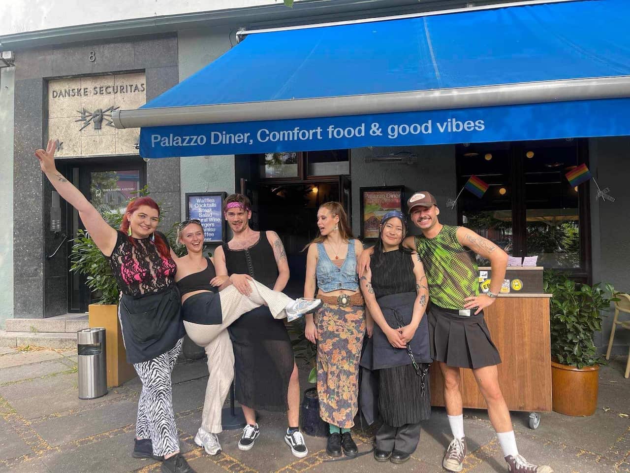 A group of friends stands in front of the pub with rainbow flags.